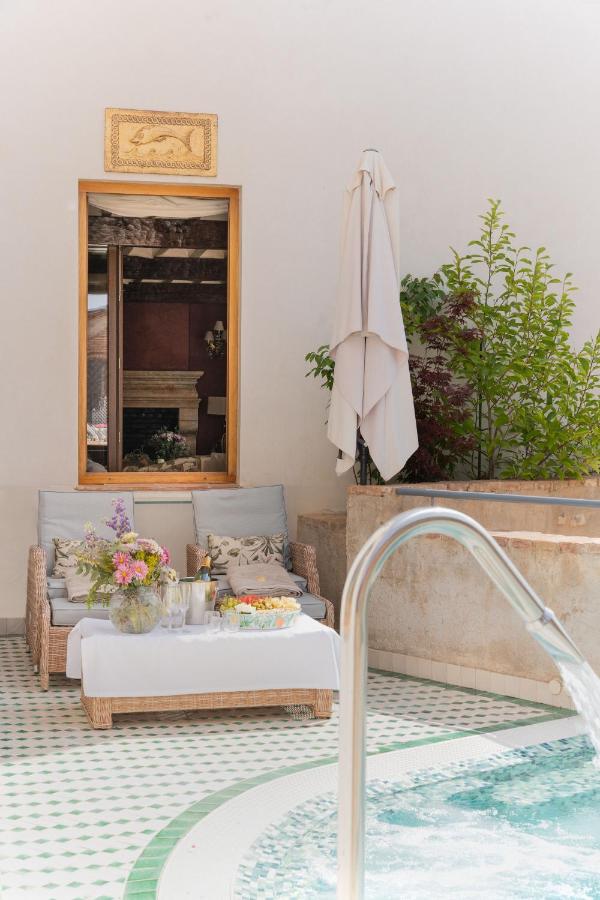 A cozy outdoor seating area, reminiscent of boutique hotels in Granada, features a wicker sofa, a table with flowers, drinks, and snacks by a small pool. Lush plants and a closed white umbrella sit against a cream wall topped with a window.
