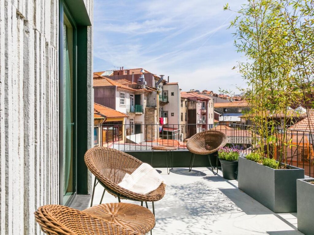 Top 12 Boutique Hotels in Porto, Portugal 20 A modern balcony with wicker chairs and potted plants, reminiscent of boutique hotels in Porto, overlooks a charming cityscape of red-roofed buildings under a partly cloudy sky.