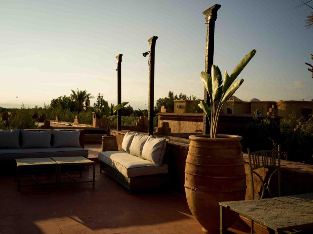 10 Unique Riads in Marrakech with Pool 21 Outdoor patio with cushioned seating, large potted plants, and rustic decor in warm sunlight—reminiscent of riads in Marrakech with pool touches. Masonry and wooden elements blend with lush greenery and buildings in the background.