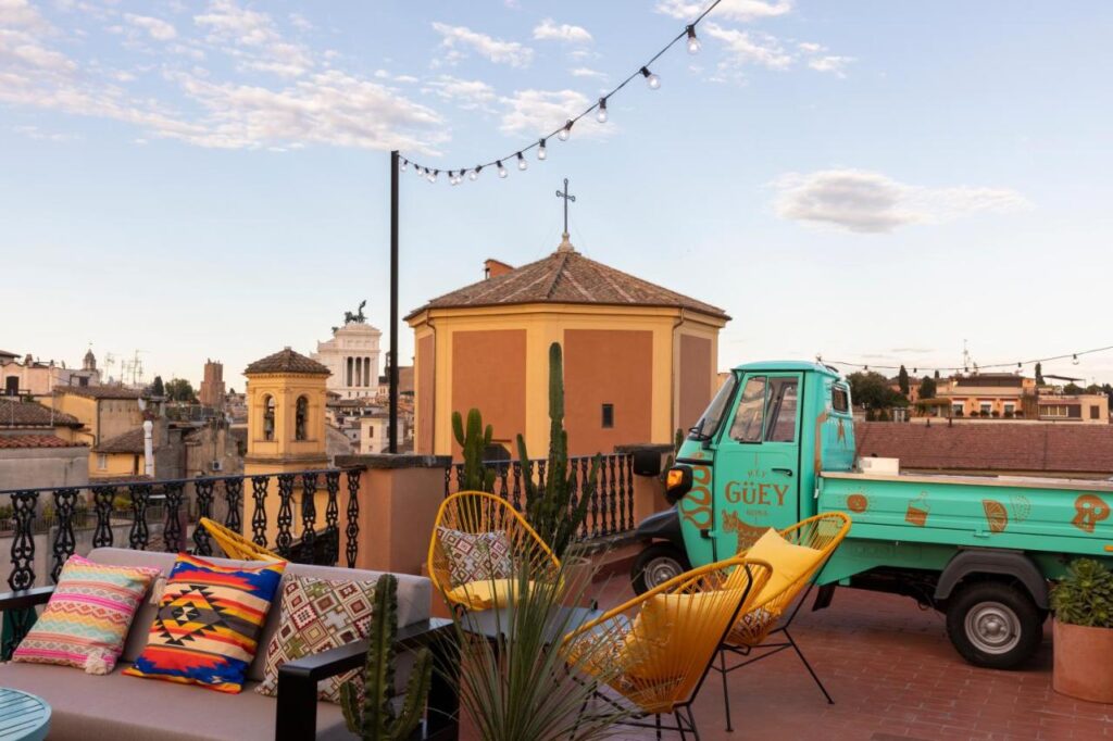 13 UNIQUE Boutique Hotels in Rome 8 Colorful rooftop terrace with patterned pillows, yellow chairs, and potted cacti, overlooking city buildings—perfect inspiration for boutique hotels in Rome Italy. A turquoise food truck sits under string lights near a round yellow building and cross.