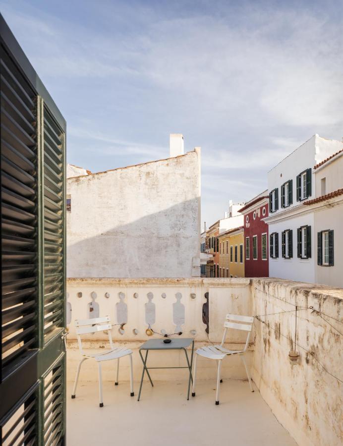 Unique Boutique Hotels in Menorca 17 A small balcony at one of the boutique hotels in Menorca features two white chairs and a round table, overlooking neighboring buildings with colorful facades and shuttered windows under a partly cloudy sky.