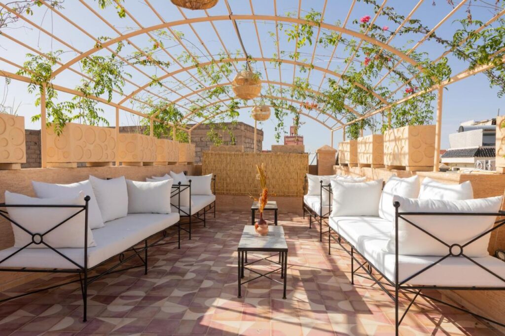 10 Unique Riads in Marrakech with Pool 25 Rooftop terrace with white cushioned sofas, a small central table, and a pergola covered with green vines and hanging lights. The patterned tile flooring adds charm, reminiscent of riads in Marrakech with pool, creating a sunny, open atmosphere.