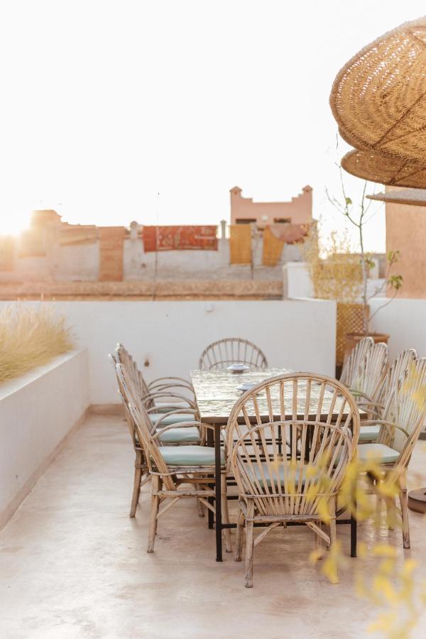 10 Unique Riads in Marrakech with Pool 12 A sunlit rooftop patio with a long table and eight wicker chairs evokes the relaxed elegance of riads in Marrakech with pool, featuring white walls, beige floors, and a cityscape of warm-toned buildings in the background.