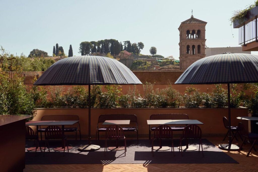 13 UNIQUE Boutique Hotels in Rome 17 Outdoor terrace with tables and chairs under dome-shaped umbrellas, overlooking greenery and a historic stone bell tower—a serene setting reminiscent of boutique hotels in Rome, Italy. Trees and classic buildings complete the sunny day backdrop.