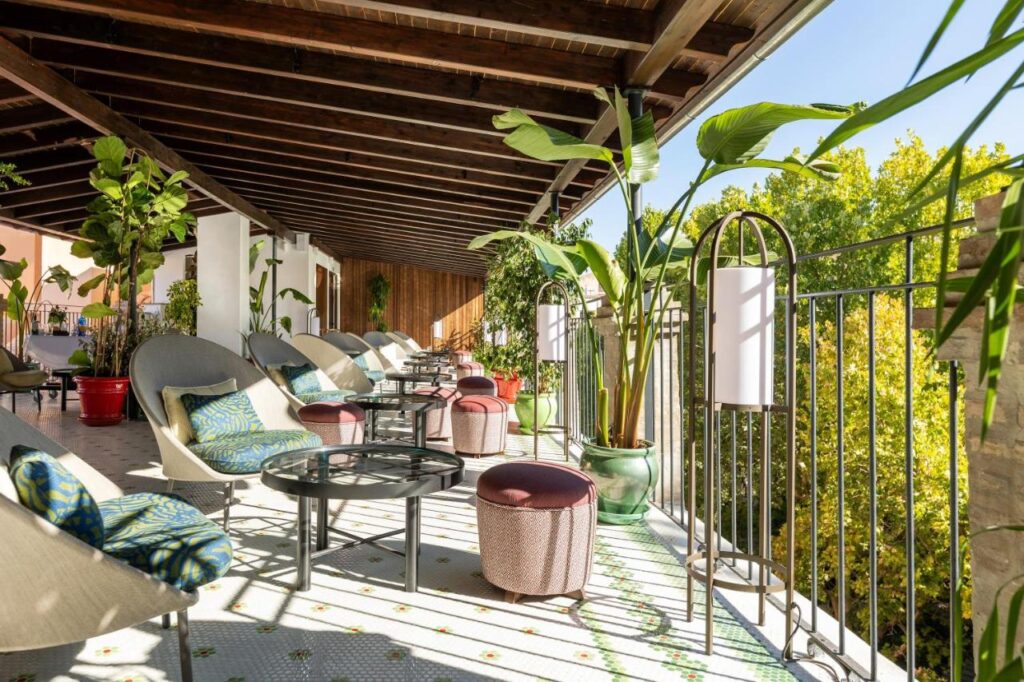 A sunny, covered terrace at one of the boutique hotels in Granada features cushioned lounge chairs, small tables, colorful stools, potted plants, and white lamps, with views of lush green trees beyond the metal railing.