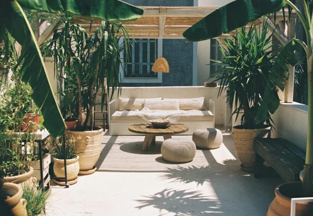 A cozy outdoor patio reminiscent of the best boutique hotels in Athens, with a shaded pergola, potted plants, cushioned seating, a round wooden table, two poufs, and sunlight casting gentle shadows on the floor.