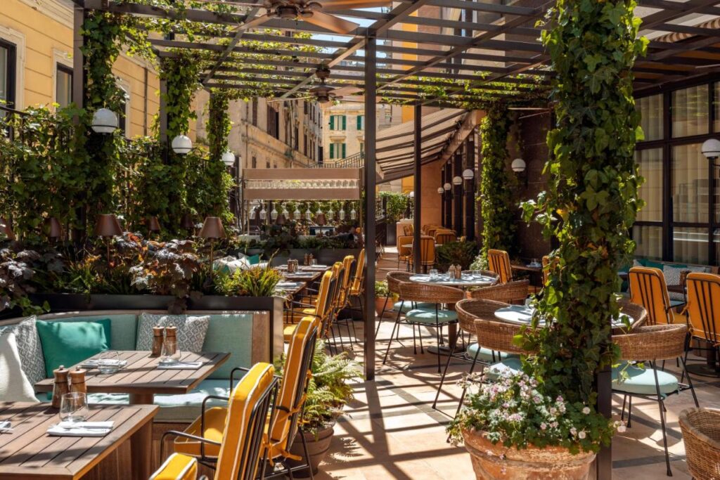 13 UNIQUE Boutique Hotels in Rome 25 Outdoor restaurant patio with tables and chairs, surrounded by green plants and vines climbing wooden pergolas. Sunlight casts shadows on the tiled floor, creating a cozy ambiance reminiscent of boutique hotels in Rome, Italy.