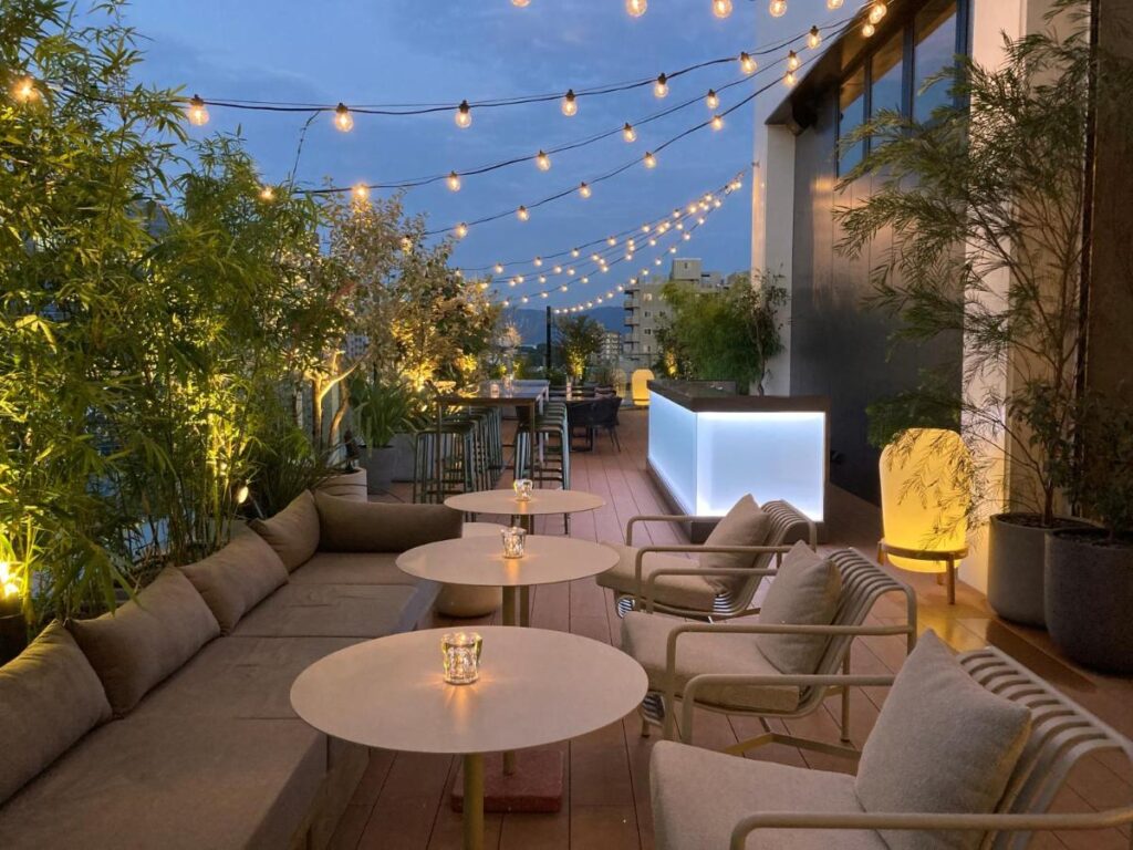 7 Best Hotels in Hiroshima, Japan 11 A rooftop terrace at dusk with string lights, cozy seating, round tables, potted plants, and a glowing modern bar—this inviting space rivals the best hotels in Hiroshima Japan for relaxing or socializing.