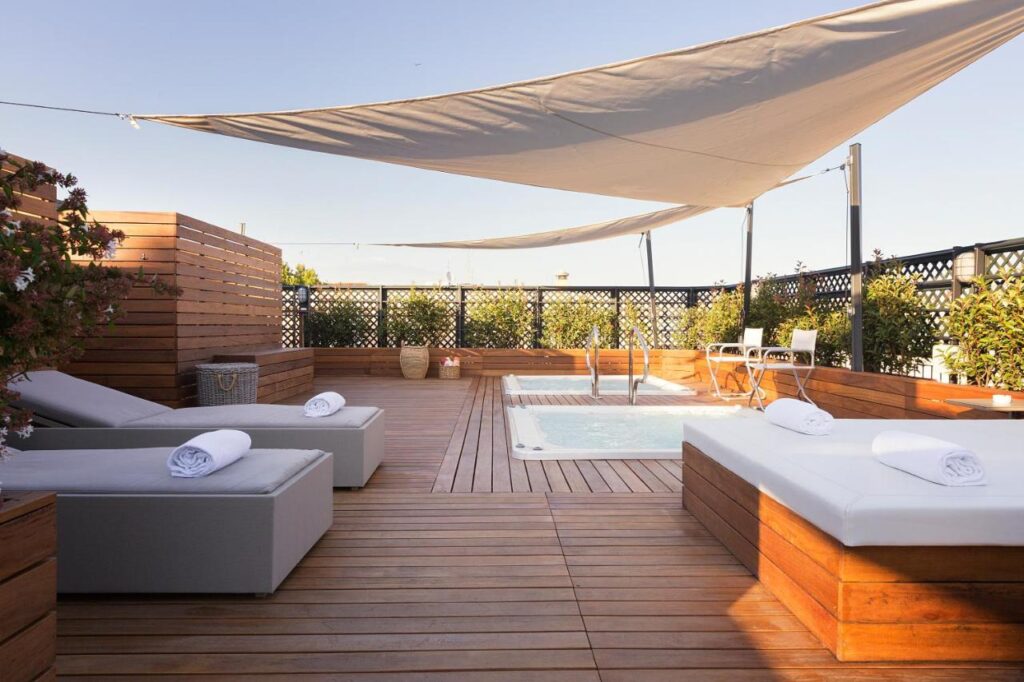 13 UNIQUE Boutique Hotels in Rome 22 A rooftop deck at one of the boutique hotels in Rome, Italy features wooden flooring, a small pool, white cushioned lounge chairs with rolled towels, potted plants, a shade sail overhead, and a small table with chairs in the background.