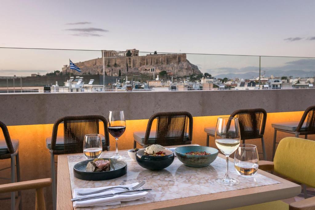 A rooftop dining table with wine and Mediterranean dishes overlooks the Acropolis in Athens at sunset, capturing the elegant ambiance found at boutique hotels in Athens, with modern chairs and a cityscape visible in the background.