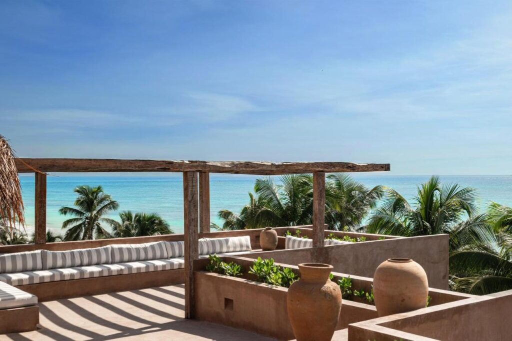Top Hotels in Tulum, Mexico: Luxury and Beachfront Stays 5 A rooftop terrace at one of the top hotels in Tulum features striped cushioned seating, clay pots, and a wooden pergola overlooking palm trees and a turquoise ocean beneath a clear blue sky.