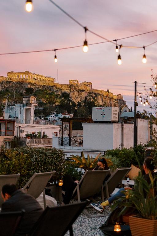 People relax on a rooftop patio at sunset, with string lights overhead and plants around. In the distance, the Acropolis and Parthenon glow—an iconic view from many boutique hotels in Athens under a colorful sky.