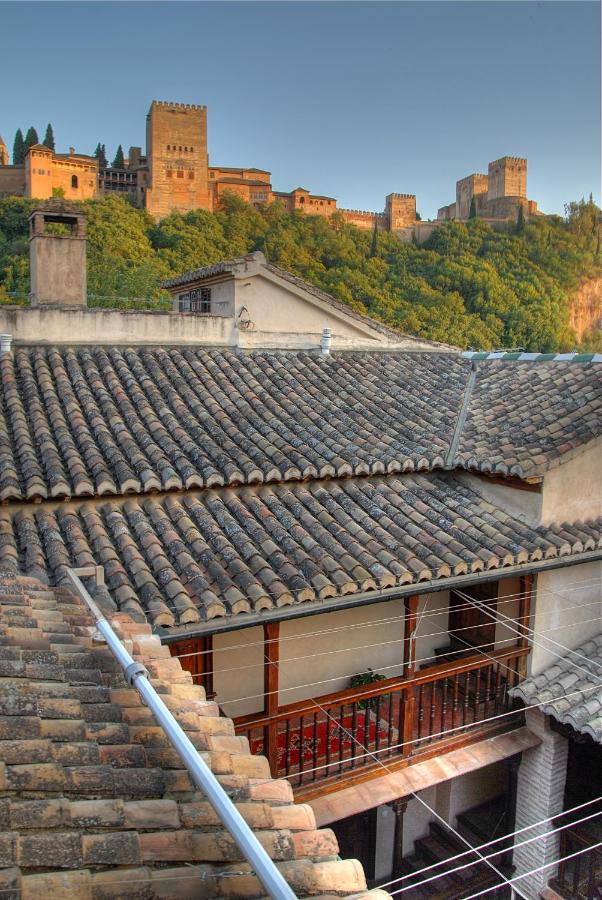 Rooftops of traditional Spanish houses with tiled roofs in the foreground, and the Alhambra palace complex—near many boutique hotels in Granada—on a lush green hilltop in the background under a clear sky.