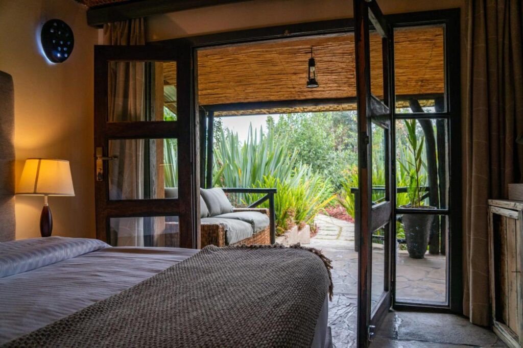 Best Hotels in Rwanda: Unique Hotels and Lodges 2 A cozy bedroom in one of the best hotels in Rwanda, featuring a neatly made bed, a lit lamp, and open glass doors leading to a patio with a couch, overlooking lush green plants and a stone path in a garden setting.