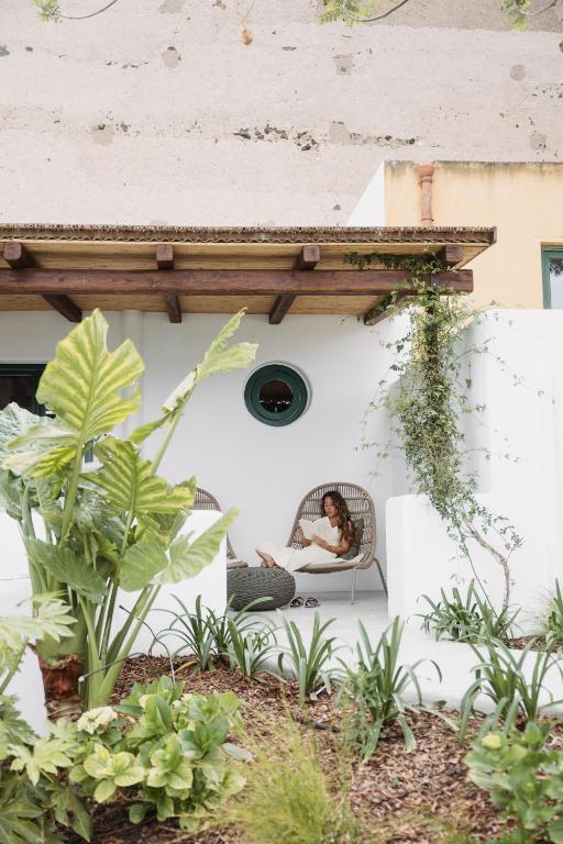 7 Best Hotels in the Aeolian Islands 14 A woman relaxes in a wicker chair on a white patio surrounded by lush green plants, beneath a rustic wooden roof—an inviting scene often found at charming hotels in the Aeolian Islands. She reads, enjoying the peaceful outdoor setting.