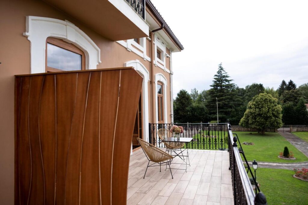 Balcony with two modern chairs and a small table overlooks a garden and trees, attached to a beige building—similar to stylish hotels in Cantabria—with large windows and white trim on a cloudy day.