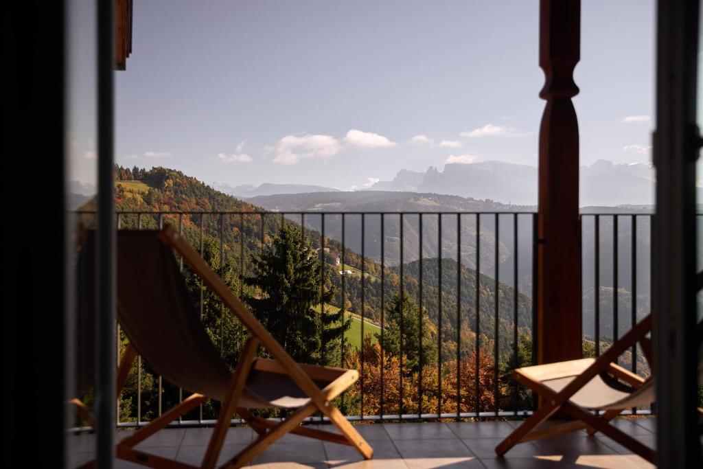 A balcony with two wooden lounge chairs overlooks a scenic mountain landscape, reminiscent of views found at the best hotels in the Dolomites, with rolling hills, green forests, and distant peaks under a clear sky.