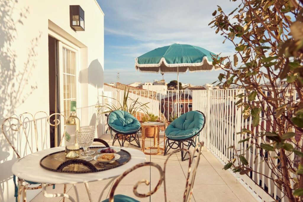 Unique Boutique Hotels in Menorca 8 A sunny balcony like those in boutique hotels in Menorca, with two teal cushioned chairs, a round table set with drinks and snacks, potted plants, and a teal umbrella. White railings and a cityscape complete the stylish scene.