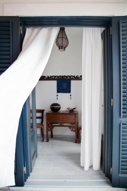 7 Best Hotels in the Aeolian Islands 22 View through open blue shutters and flowing white curtains into a small, bright room—reminiscent of charming hotels in the Aeolian Islands—with a wooden table, two chairs, wall art, a black vase, and a hanging light fixture above.
