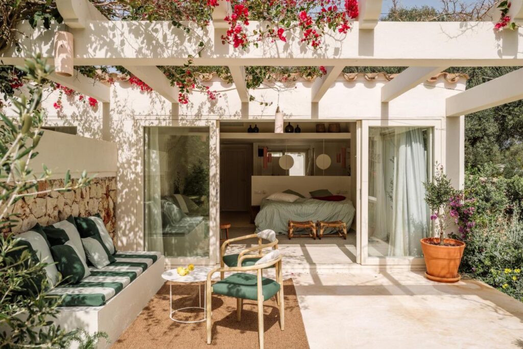 Unique Boutique Hotels in Menorca 10 A sunlit patio with a cushioned bench, a chair, and a small table overlooks an inviting bedroom through open glass doors—just the kind of cozy outdoor space you’ll find at charming boutique hotels in Menorca, with flowering vines trailing from a white pergola above.
