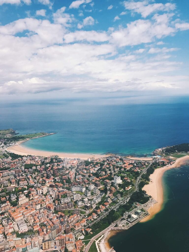 Aerial view of a coastal city with clusters of buildings, sandy beaches, green spaces, blue ocean, and several hotels in Cantabria under a partly cloudy sky.