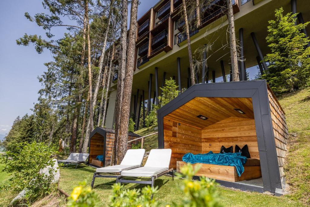 Two modern wooden cabanas with cozy seating and pillows sit on a grassy slope beneath a stilted building among tall trees, evoking the charm of the best hotels in the Dolomites, with two lounge chairs arranged invitingly on the lawn.