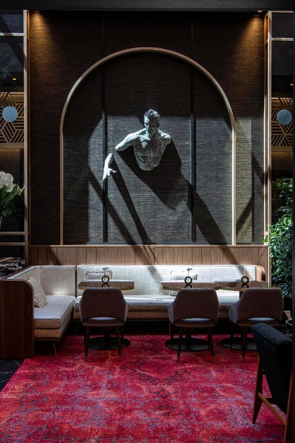 13 UNIQUE Boutique Hotels in Rome 6 A modern lounge area in the style of boutique hotels in Rome, Italy, with a red rug, cushioned seating, and a wall-mounted sculpture of a man’s upper body emerging from a dark arched alcove. Sunlight casts dramatic shadows across the space.