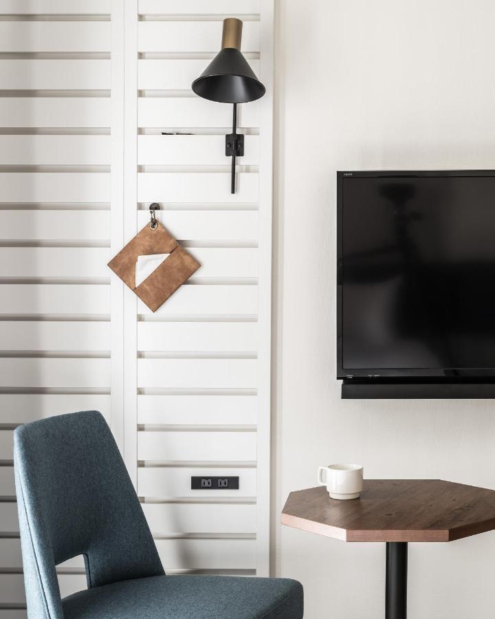 7 Best Hotels in Hiroshima, Japan 16 A modern interior evokes the style of the best hotels in Hiroshima, Japan, featuring a blue chair, hexagonal wooden table with a white mug, wall-mounted TV, black wall lamp, and a decorative leather keychain on a white slatted wall.
