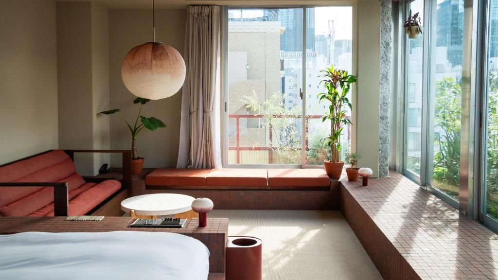 15 Unique Hotels in Tokyo: Coolest hotels 38 A modern, sunlit hotel room with large windows, red-brown seating, potted plants, a white bed, and a round hanging lamp; city buildings are visible outside—perfect for those seeking unique hotels in Tokyo.