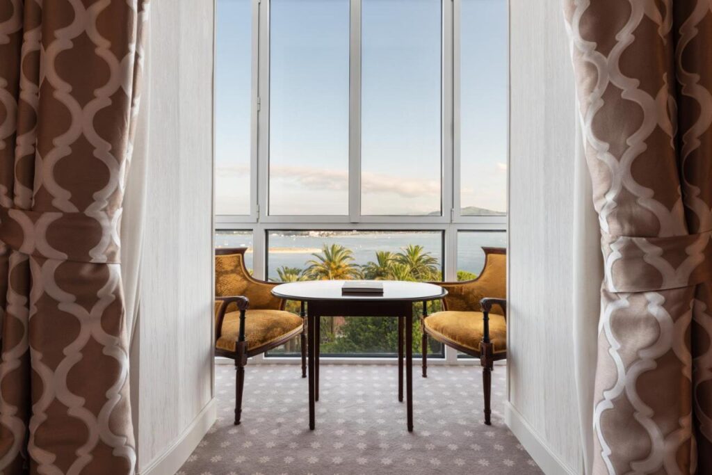 A round table with two vintage upholstered chairs sits by tall windows, overlooking palm trees and a body of water—an inviting scene reminiscent of charming hotels in Cantabria. Patterned curtains frame the cozy seating area, creating a relaxing atmosphere.