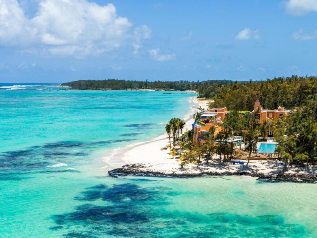 Most Unique Adults Only Hotels in Mauritius, Indian Ocean 7 A tropical beach with clear turquoise water, white sand, and lush green trees. Colorful buildings and a pool from adults only hotels in Mauritius line the shoreline under a blue sky with scattered clouds.