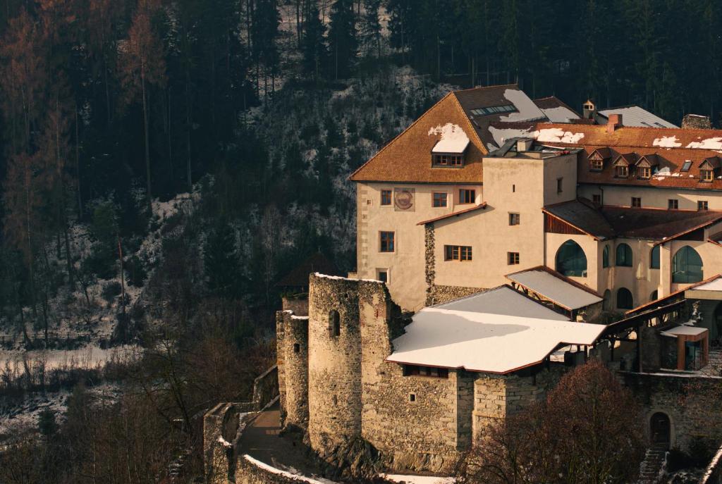 A historic castle with stone walls and snow-covered roofs sits on a forested hillside—like one of the best hotels in the Dolomites—surrounded by trees dusted with snow under soft, golden light.