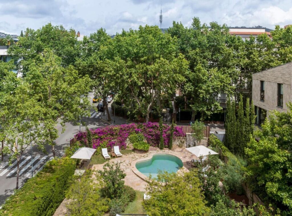 13 Unique Hotels in Barcelona, Spain 13 A small, heart-shaped pool is surrounded by lounge chairs and umbrellas in a lush garden with blooming purple flowers—one of the charming features found at unique hotels in Barcelona, bordered by trees and nearby residential buildings along a quiet street.