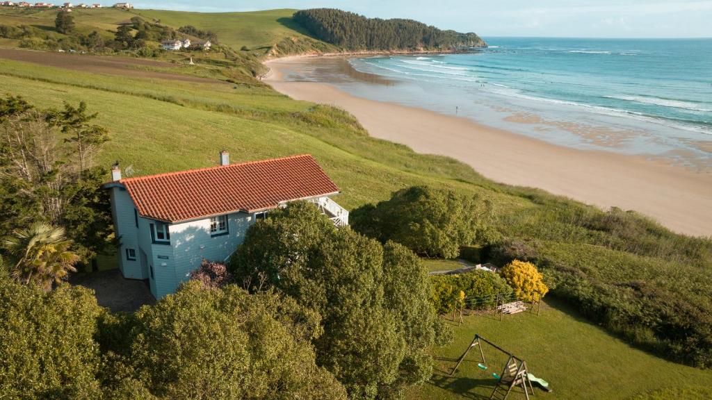 A coastal house with a red-tiled roof sits among lush greenery near a sandy beach—perfect for those seeking tranquil hotels in Cantabria—with gentle waves, green hills, and a charming swing set in the yard.