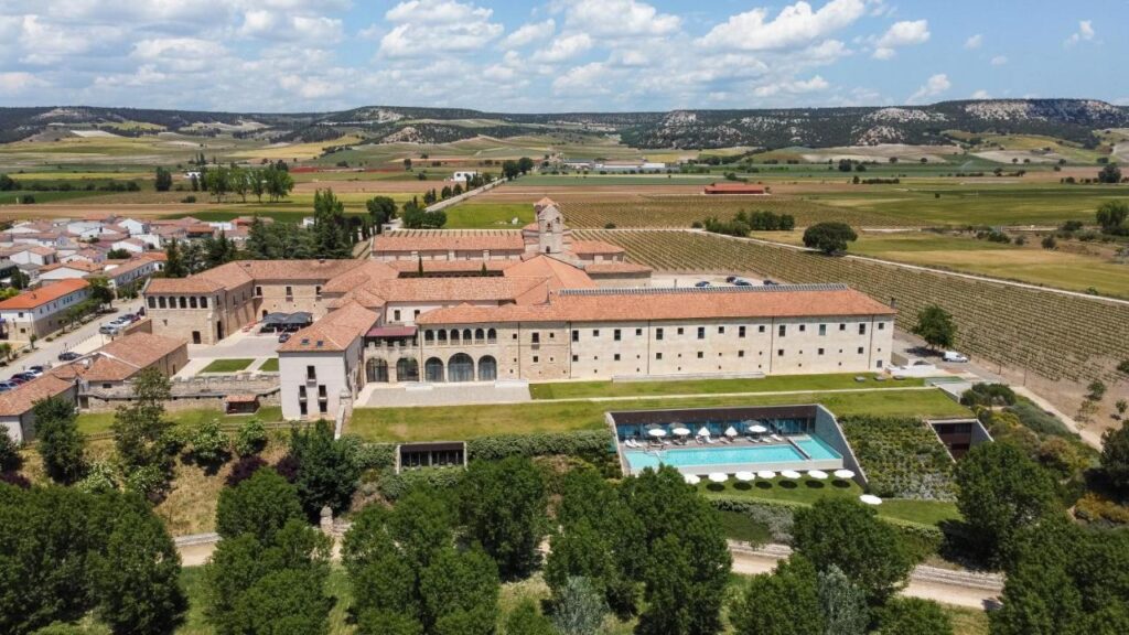 4 Best Hotels in Ribera del Duero, Spain 18 Aerial view of a large historic building with red-tiled roofs surrounded by vineyards and hills, featuring a rectangular outdoor pool and manicured gardens—an idyllic example of luxury hotels in Ribera del Duero.