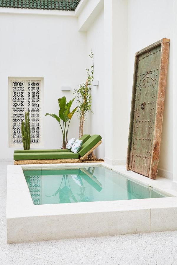 10 Unique Riads in Marrakech with Pool 11 A small, square pool in a bright white courtyard reflects the serene charm of riads in Marrakech with pool, complete with a green cushioned lounge chair, potted plants, and a large rustic wooden door leaning against the wall.
