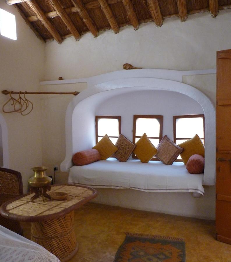 Authentic Heritage Hotels in Mandawa 6 A cozy alcove with small windows, a built-in white bench topped with colorful cushions, rustic wooden furniture, and a woven rug—all under a thatched ceiling inspired by the charming hotels in Mandawa—creates a warm, inviting room.