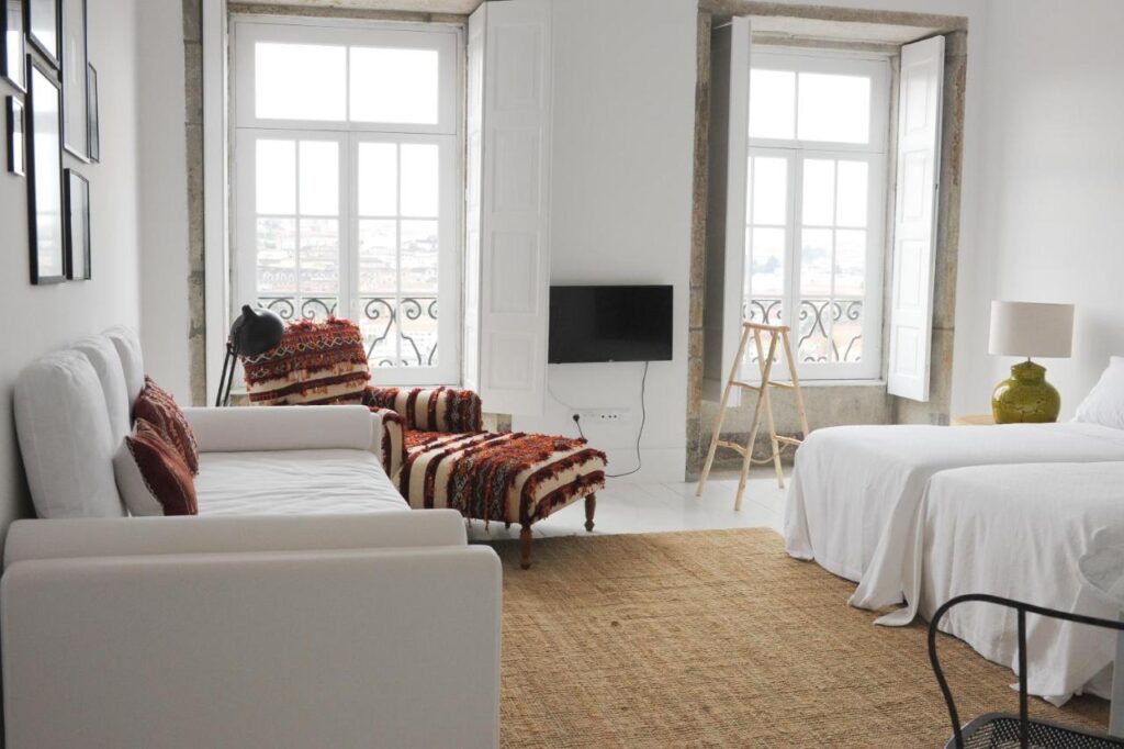 Top 12 Boutique Hotels in Porto, Portugal 46 Bright, minimalist bedroom reminiscent of boutique hotels in Porto, featuring two large windows, white walls, a bed, sleek sofa, patterned chaise lounge, green lamp, TV, and a natural fiber rug.