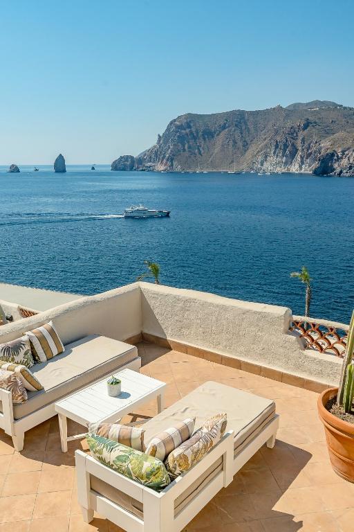7 Best Hotels in the Aeolian Islands 19 Sunny terrace with cushioned lounge chairs and table overlooking a calm blue sea, rocky cliffs, and a boat sailing in the distance—an idyllic retreat typical of hotels in the Aeolian Islands.