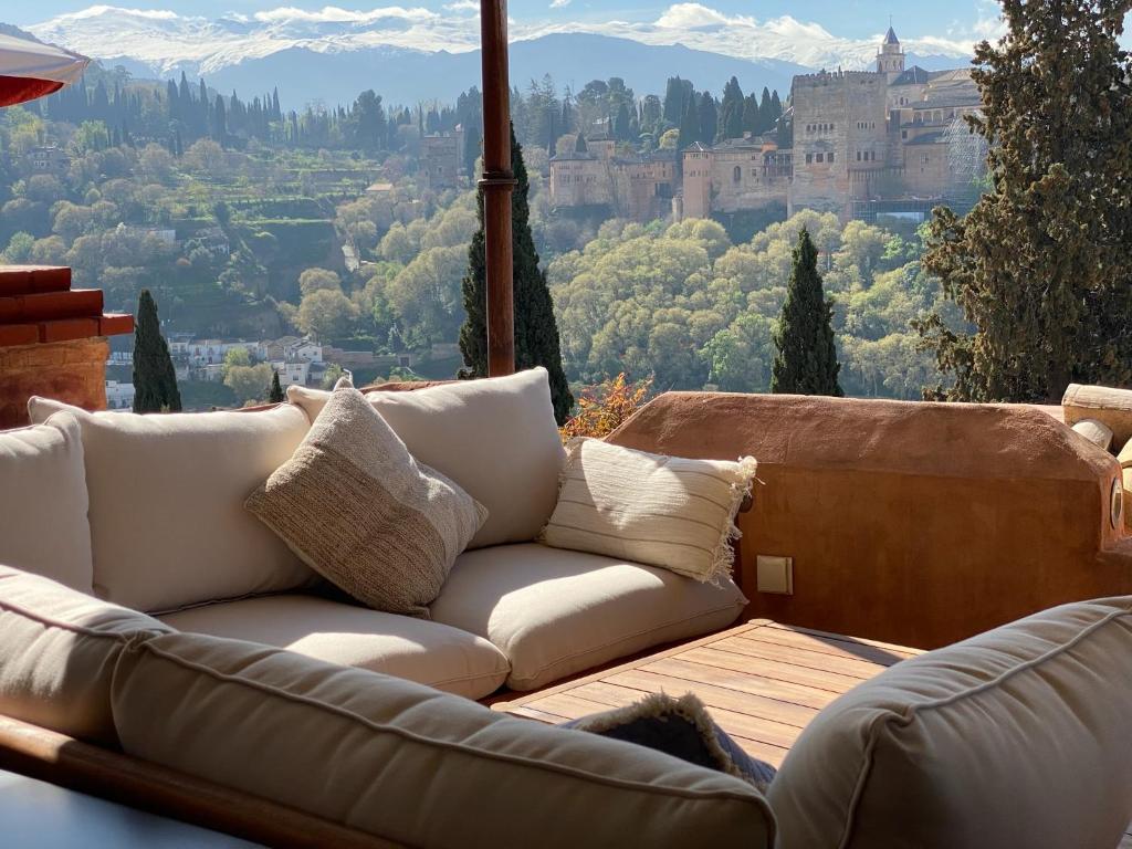 A cozy outdoor sofa with beige cushions sits on a wooden terrace, overlooking the Alhambra palace and lush greenery—an inviting scene often found at boutique hotels in Granada, with snow-capped mountains under a clear sky.