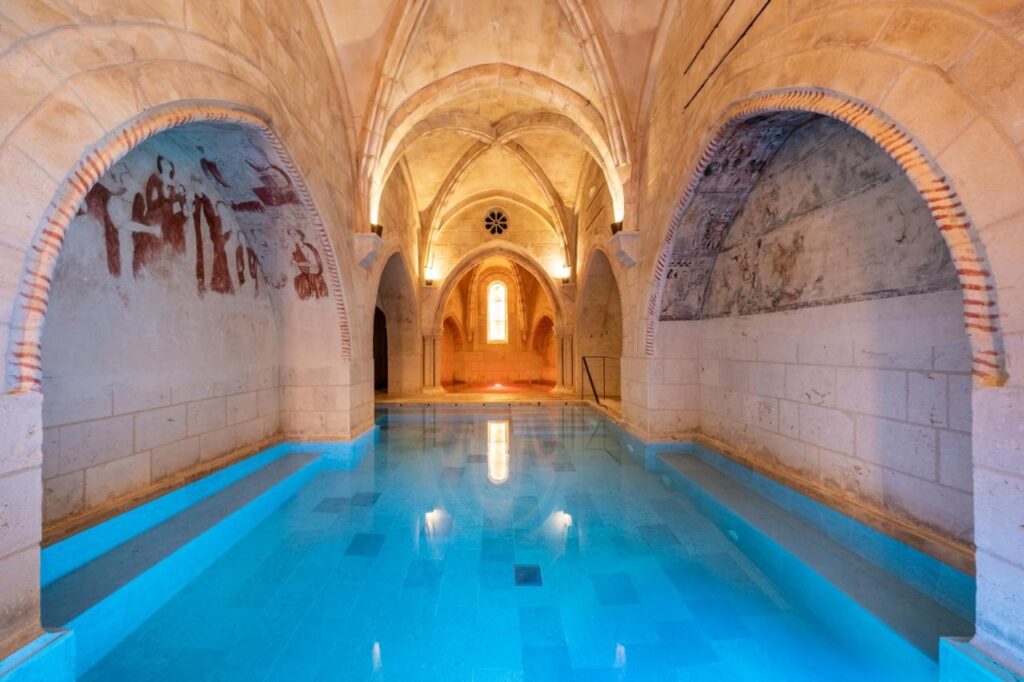 4 Best Hotels in Ribera del Duero, Spain 19 A serene indoor pool with bright blue water sits beneath arched stone ceilings and walls, much like those found in exclusive hotels in Ribera del Duero, featuring faded murals and soft natural light streaming through a distant arched window.