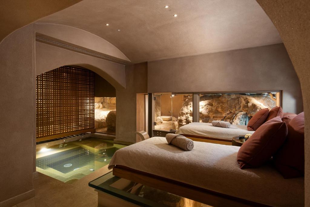 A luxurious spa room in one of the finest boutique hotels in Athens, featuring two massage beds, rolled towels, soft lighting, and a small indoor pool. Large windows reveal a rocky, cave-like feature for a serene atmosphere.