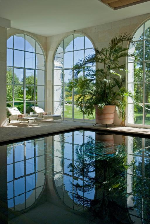 TOP 10 Luxury Hotels in Andalucía 7 Indoor pool with clear water reflecting large arched windows, two lounge chairs, and a tall potted palm. Sunlight streams in, revealing green trees outside—evoking the bright, luxurious ambiance found in top luxury hotels in Andalucía.