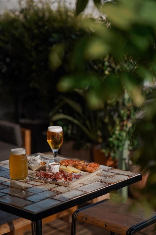 13 Unique Hotels in Barcelona, Spain 29 A rustic outdoor table set with cured meats, cheese, bread, a glass of beer, and a mason jar with a yellow drink evokes the relaxed charm found at unique hotels in Barcelona, surrounded by lush greenery and blurred foliage in the foreground.