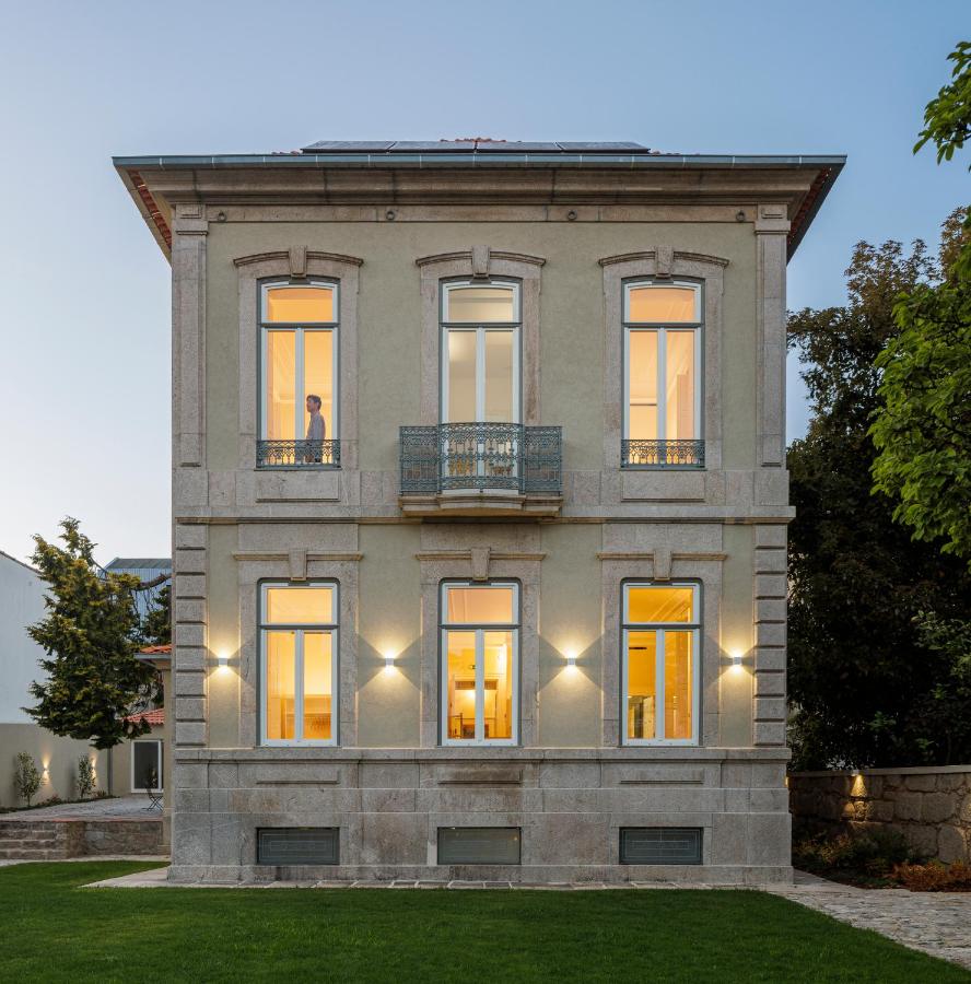 Top 12 Boutique Hotels in Porto, Portugal 13 A two-story beige house with large, illuminated windows at dusk evokes the charm of boutique hotels in Porto. A person stands inside an upper window; the home features a small balcony, stone base, and is surrounded by green lawn and trees.