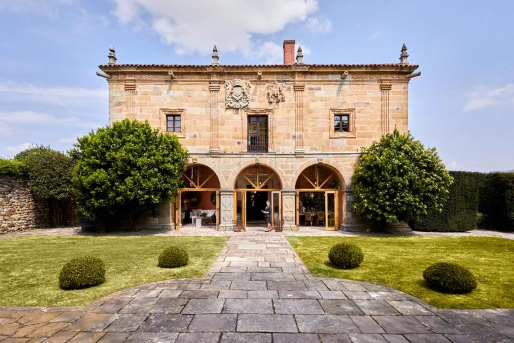 A stone mansion with arched windows and decorative carvings stands behind a patio with neatly trimmed bushes and two symmetrical trees—reminiscent of the elegance found at luxury hotels in Cantabria—set under a blue sky with scattered clouds.