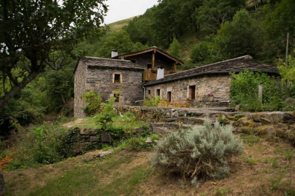 A rustic stone house with a wooden roof sits surrounded by greenery, trees, and wildflowers on a grassy hillside—reminiscent of charming hotels in Cantabria. The peaceful rural setting features gardens and terraced stone walls under an overcast sky.