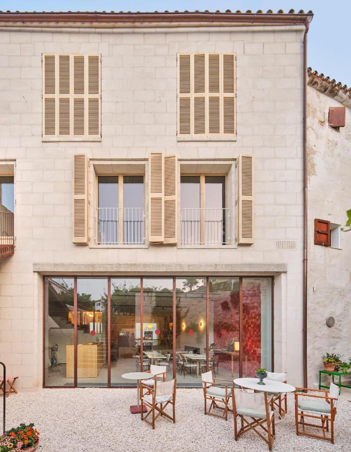 Unique Boutique Hotels in Menorca 34 A modern stone building typical of boutique hotels in Menorca, featuring large glass doors, wooden shutters, an outdoor seating area on a gravel patio with potted flowers, and warm indoor lighting visible through the windows.