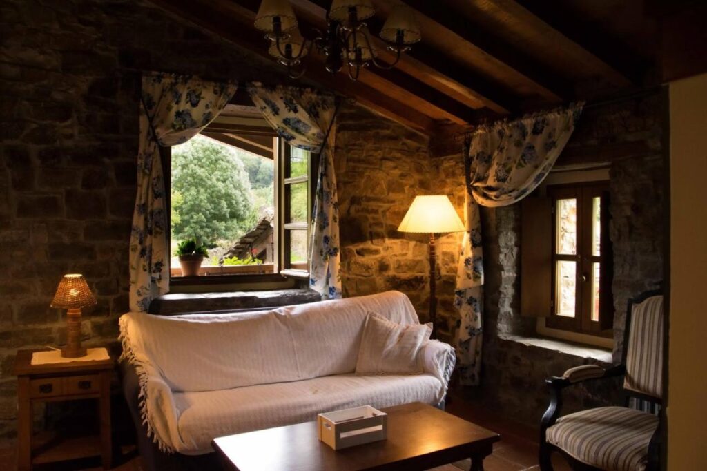 Cozy rustic living room with stone walls, a white-covered sofa, wooden ceiling beams, floral curtains, and warm lamp lighting—evoking the inviting charm found in boutique hotels in Cantabria. Green trees are visible outside the main window.