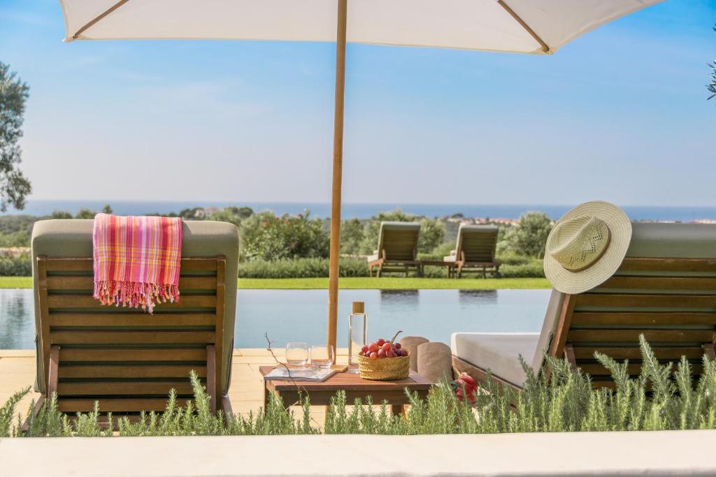 Unique Boutique Hotels in Menorca 40 Two lounge chairs face a pool and distant sea view under a large umbrella at one of the boutique hotels in Menorca. A hat, colorful towel, drinks, and fruit basket rest nearby, creating a relaxing summer atmosphere under the clear, sunny sky.